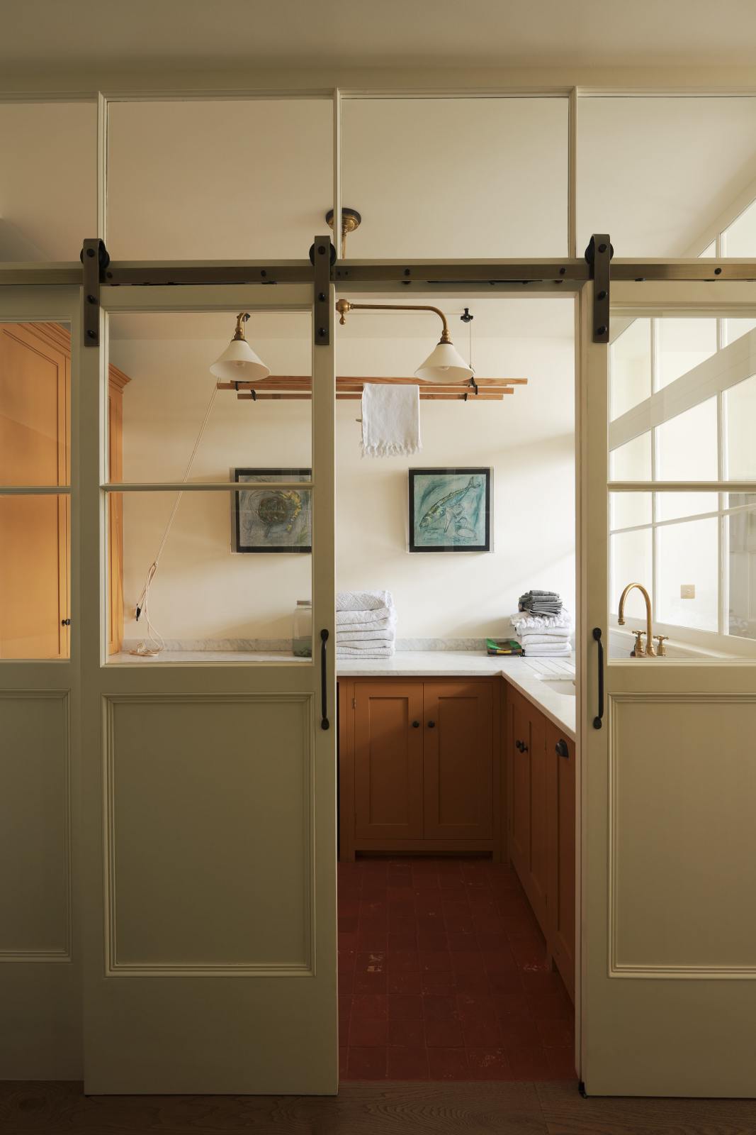 A cream sliding door leads to a scullery yellow laundry room featuring an L-shaped sink with a white marble countertop and a stunning aged brass Ionian faucet. Neatly stacked white towels are arranged in layers, and two framed artworks adorn the walls. In the centre of the ceiling hangs a deVOL Gaselier light, while a deVOL Laundry Maid, with a towel draped over it, is positioned behind.