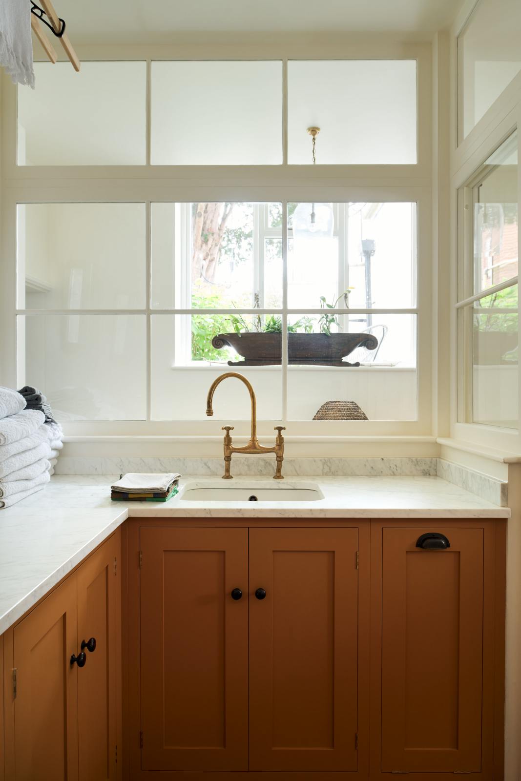 A closer look at a scullery yellow laundry room reveals an L-shaped sink with a white marble countertop and a stunning aged brass Ionian faucet. Neatly stacked white towels are arranged in layers, and the view through the window lends the space a beautiful sense of calm.