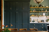 The spacious kitchen in the renovated rectory features a blue L-shaped countertop, a mix of cabinetry against green walls, and a metro tile backsplash. At the centre, a copper-topped table flanked by a vintage bench and chairs is complemented by an elegant antique chandelier.