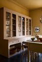 An angled view of a warm yellow room features a large pink dresser, showcasing glazed cupboards filled with delicate ceramics, papier-mâché mushrooms, and wooden baskets at the bottom, creating a relaxed, living-room atmosphere. The space includes a wide prep table with an open nook for deVOL bum stools, large ceramic bowls, a metal vines-and-flowers chandelier, and a golden-framed painting of a fish and a lemon.