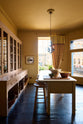 A warm yellow room with a large Princelet pink dresser showcasing glazed cupboards filled with delicate ceramics, papier-mâché mushrooms, and wooden baskets along the bottom, giving the space a relaxed, living-room feel. The space includes a wide prep table with an open nook for deVOL bum stools. There are large ceramic bowls, a metal vines-and-flowers chandelier, and a golden-framed painting of a fish and a lemon.