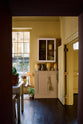 The room is painted a warm yellow and features a wide prep table with an open nook for deVOL bum stools. A Princelet pink base cupboard adds a cheerful touch, while a vintage white metal wall cupboard provides additional storage. The space is beautifully decorated with mustard yellow accents, vintage ceramic vases and furniture, and a plate illustrated with citrus fruits. A vintage gold-finish metal side table completes the bohemian style of the room.