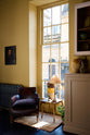 This room features an angled view of a warm yellow cupboard with a Princelet pink base beside a tall window, along with a vintage white metal wall cupboard. The space is lovingly decorated with mustard yellow accents, vintage ceramic vases, and furniture, including a small oak brown armchair. An oil portrait painting adorns the wall, complemented by a vintage gold-finish metal side table, all contributing to a bohemian style.