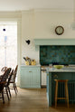 A corner of a humble and calming turquoise, classic English kitchen featuring a cooker and sink run with a Carrara marble worktop, a suspended false chimney breast, a glazed wall cupboard, and a kitchen island with Carrara marble on one side and solid oak on the other. Adorned with symmetrical hanging lights, butter-yellow ceramics, deVOL bum stools and ocean porcelain tiles, it creates a warm and simple home kitchen.