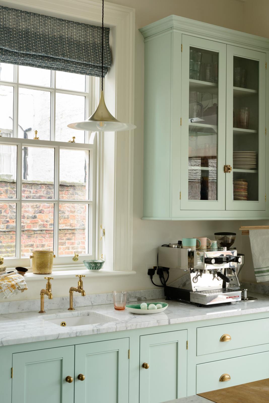 A corner of a humble and calming turquoise, classic English kitchen featuring a cooker and sink run with a Carrara marble worktop, a suspended false chimney breast, a glazed wall cupboard, and a kitchen island with a solid oak worktop. Adorned with symmetrical hanging lights, artisan ceramics, silver coffee machines and fresh eggs, it creates a warm and simple home kitchen.