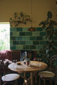 A quiet corner of a chic and glamorous Bobbin Black Haberdasher’s bar featuring vineyard tables, bentwood café chairs, petite amber frilly glass pendant lights, and a large display of Emerald green London splashback tiles. Adorned with a long antique couch, plants, vintage watering cans, and deVOL candles, it creates a beautiful and intimate personal bar.