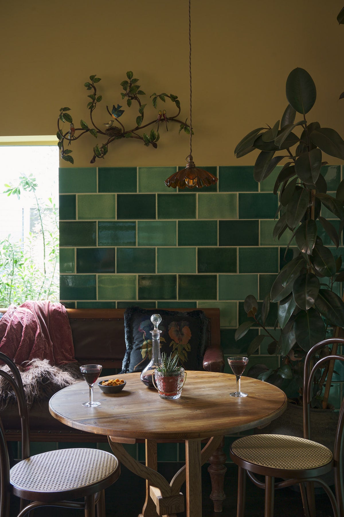 A quiet corner of a chic and glamorous Bobbin Black Haberdasher’s bar featuring vineyard tables, bentwood café chairs, petite amber frilly glass pendant lights, and a large display of Emerald green London splashback tiles. Adorned with a long antique couch, plants, vintage watering cans, and deVOL candles, it creates a beautiful and intimate personal bar.
