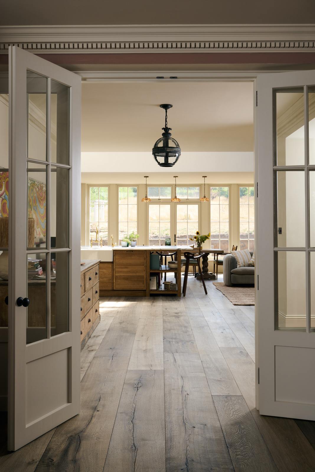 Past white French doors is an elegant and spacious Haberdasher’s kitchen features a large cooker run, a kitchen island with a double Belfast sink, an aged brass Ionian tap, an Arabescato marble worktop, splashback, and glazed cupboards. Adorned with vintage furniture, artisan ceramics, glass flower pendant lights, and a sunflower arrangement, this kitchen creates a classic social space with functional storage.