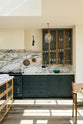A quiet corner of an elegant and spacious Haberdasher’s kitchen features a large cooker run, a kitchen island with an Arabescato marble worktop, a splashback, and glazed cupboards. Adorned with vintage furniture, artisan ceramics, glass pendant lights, copper pans, and a bowl of lemons, this kitchen creates a classic social space with functional storage.