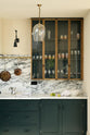 A quiet corner of an elegant and spacious Haberdasher’s kitchen features a large cooker run with an Arabescato marble worktop, a splashback, and glazed cupboards. Adorned with vintage items, artisan ceramics, glass pendant lights, copper pans, and limes, this kitchen creates a classic social space with functional storage.