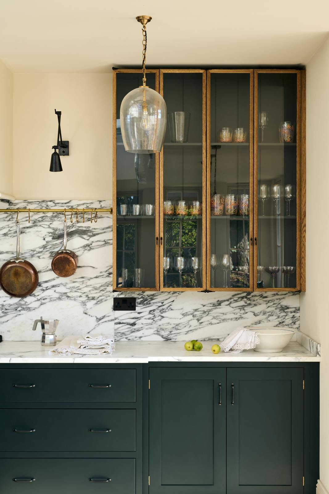 A quiet corner of an elegant and spacious Haberdasher’s kitchen features a large cooker run with an Arabescato marble worktop, a splashback, and glazed cupboards. Adorned with vintage items, artisan ceramics, glass pendant lights, copper pans, and limes, this kitchen creates a classic social space with functional storage.