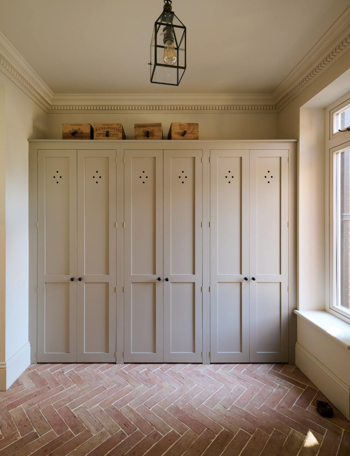 A modest foyer features bespoke storage cupboards in a muted sage green, complemented by a pale terracotta parquet floor, vintage wooden storage boxes, and a metal and glass pendant light, which adds to the coastal aesthetic.