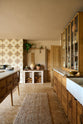 Nestled between a warm and chic Haberdasher’s kitchen features an L-shaped sink and cooker run, countertop cupboards, a kitchen island, a Carrara and copper mix worktop, and a double-fluted marble sink, is a firewood storage area. Adorned with artisan ceramics, a large vintage rug, and luscious green house plants, this kitchen creates a warm and inviting Spanish-inspired space in Yorkshire.