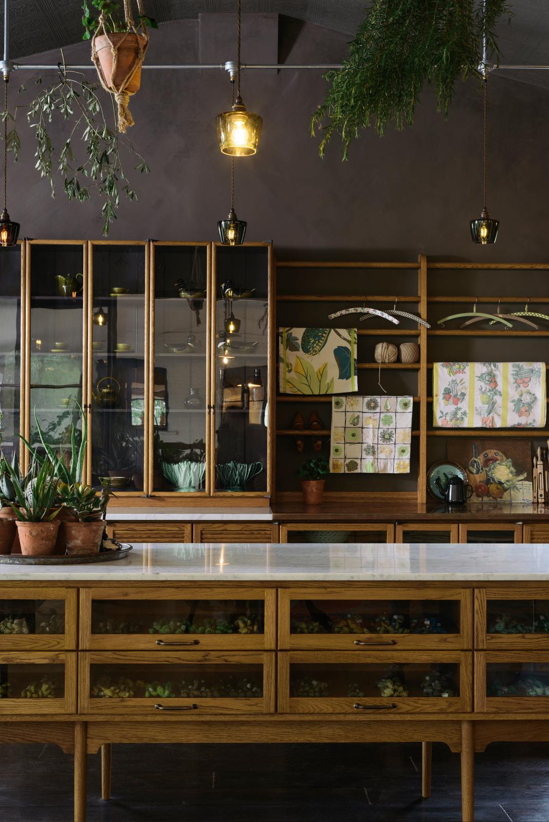 A warm and inviting Haberdasher’s kitchen features a long sink run with a Carrara marble and copper worktop, a double-fluted marble sink, a kitchen island, wall cupboards, and countertop cupboards. Adorned with luscious green hanging and potted plants, vintage items, artisan ceramics, illustrative tea towels, paintings, and a beloved white gas stove, this kitchen creates a lovely exhibitionist space that is both beautiful and practical.