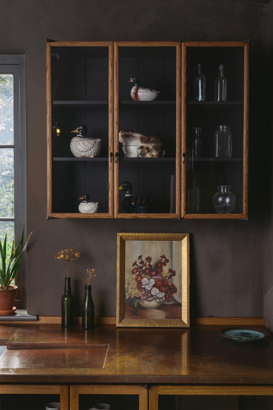 A quiet corner of a warm and inviting Haberdasher’s kitchen features a long sink run with a copper worktop, and glazed wall cupboards. Adorned with a luscious green potted plant, vintage items, artisan ceramics, and a golden framed floral painting, this kitchen creates a lovely exhibitionist space that is both beautiful and practical.
