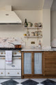 A grand and quaint Haberdasher’s kitchen features a sink and cooker run with a Paonazzo marble worktop and decorative vanity cut. Beautifully decorated with a white gas cooker with golden accents, a matching canopy, a copper pan, artisan teapot, and dried ingredients in glass jars, this kitchen creates a humble and functional space with a touch of elegance.