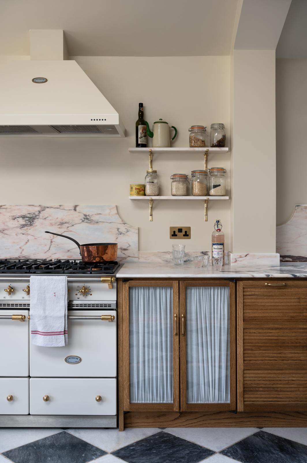 A grand and quaint Haberdasher’s kitchen features a sink and cooker run with a Paonazzo marble worktop and decorative vanity cut. Beautifully decorated with a white gas cooker with golden accents, a matching canopy, a copper pan, artisan teapot, and dried ingredients in glass jars, this kitchen creates a humble and functional space with a touch of elegance.