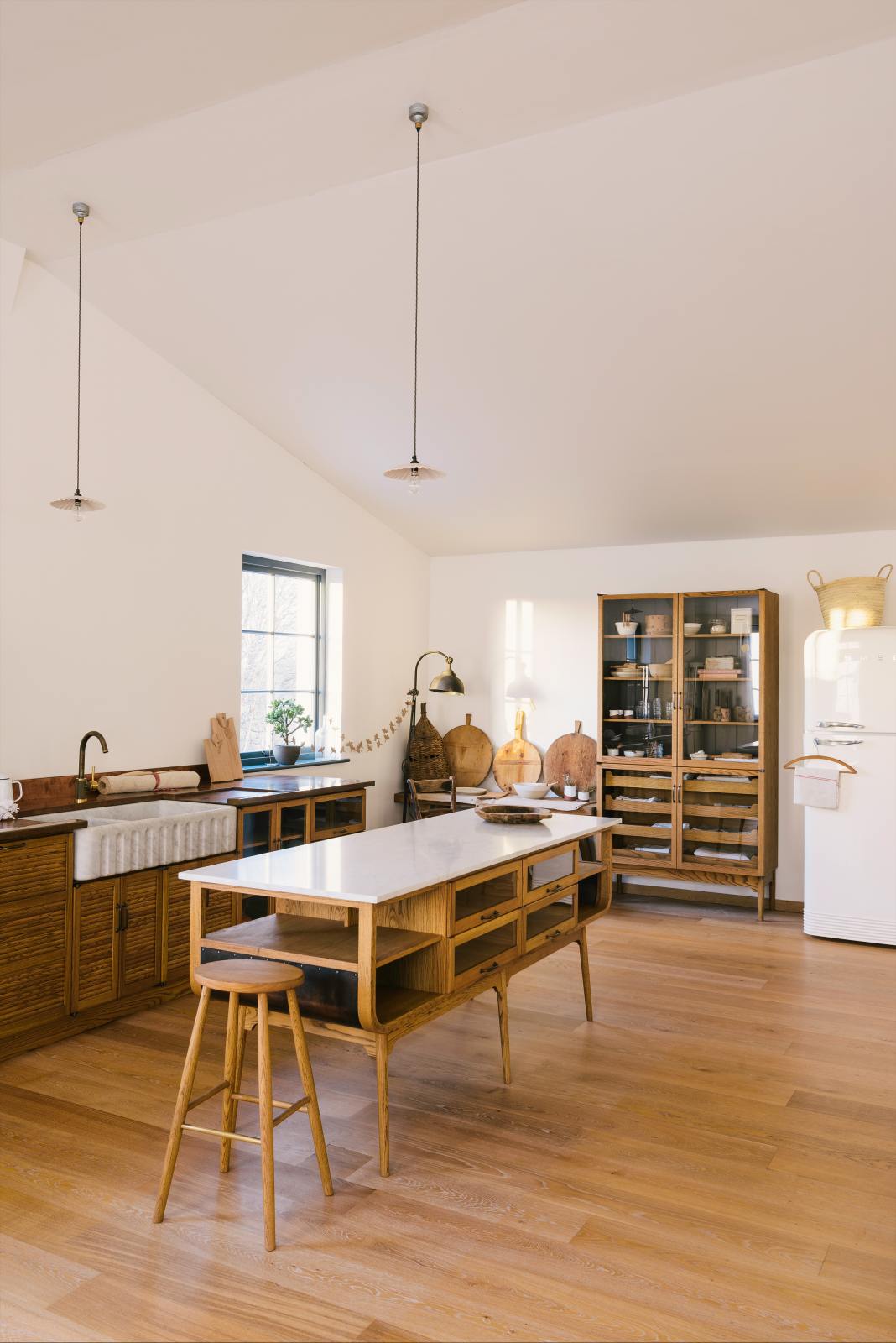 An oblique view of a warm and spacious Haberdasher’s kitchen features a sink run with a copper worktop, an Arabescato fluted sink, a small prep table with a Carrara marble countertop, and a freestanding pantry with glazed doors. Adorned with vintage furniture and items, artisan ceramics, pleat porcelain lights, large wooden tapas boards, and a delicate bird bunting in the corner of the window, this kitchen creates a contemporary yet nostalgic space.