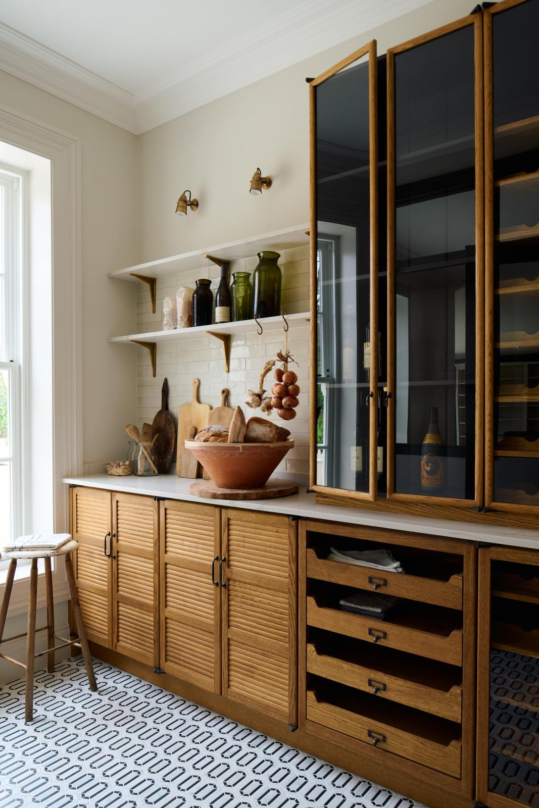 An oblique view of a smart and functional Haberdasher’s pantry features a long pantry run with glazed, open cupboards with internal drawers and tambour doors, a Carrara marble worktop and shelves, and cream splashback tiles. Beautifully decorated with eye-catching diamond mosaic flooring, oxidised brass hooks, cloves of garlic, onions, loaves of bread, and classic shelf brackets, this pantry creates a warm and neat storage space.