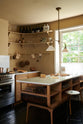 An oblique view of a cosy and sophisticated Haberdasher’s kitchen features a cooker run with a Carrara marble worktop, a small prep table, bespoke curved shelves, and muted yellow walls. Beautifully decorated with Creedy stools, a deVOL Heirloom gaselier light, aged brass hanging rails, copper pans, focaccia bread, vintage items, and artisan ceramics, this kitchen creates a bright and warm space.