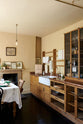 An oblique view of a cosy and sophisticated Haberdasher’s kitchen features a sink run with a copper worktop, a single marble sink, glazed countertop cupboards, and muted yellow walls. Beautifully decorated with Bentwood café chairs, vintage furniture and items, framed detailed illustrations, lace runners, tea towels, and artisan ceramics, this kitchen creates a bright and warm space.