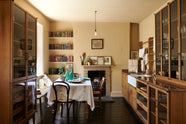 A cosy and sophisticated Haberdasher’s kitchen features a sink run with a copper worktop, a single marble sink, glazed countertop cupboards, a freestanding pantry with glazed doors, and muted yellow walls. Beautifully decorated with Bentwood café chairs, vintage furniture and items, books, framed detailed illustrations, a large platter of tapas, and artisan ceramics, this kitchen creates a bright and warm space.