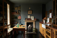 A closer look at a cosy and sophisticated Haberdasher’s kitchen features a sink run with a copper worktop, a single marble sink, glazed countertop cupboards, a freestanding pantry with glazed doors, and dark charcoal walls. Beautifully decorated with vintage furniture and items, books, framed detailed illustrations, wicker baskets, fresh bread, figs, and artisan ceramics, this kitchen creates a bright and warm space.