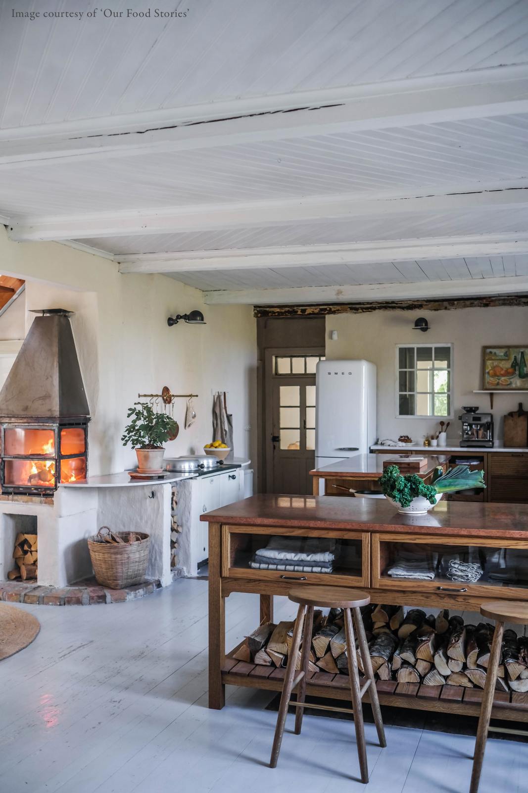 An oblique view of a warm and inviting Haberdasher’s kitchen features a sink and cooker run with a Carrara marble worktop, a double-fluted marble sink, an aged brass Ionian tap, prep tables with a copper worktop, glazed wall cupboards, and a shelf. Beautifully decorated with vintage furniture and items, artisan ceramics, firewood, a small fireplace, and a wool armchair, this kitchen creates an airy and cosy Scandinavian small fishing town hideaway.