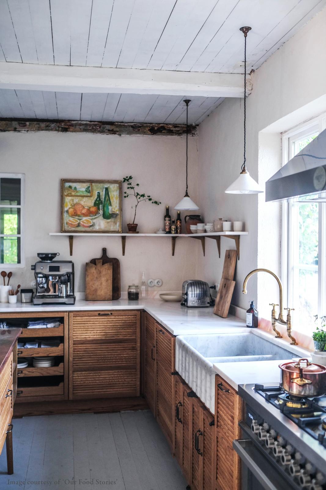 An oblique view of a warm and inviting Haberdasher’s kitchen features a sink run with a Carrara marble worktop, a double-fluted marble sink, an aged brass Ionian tap, a shelf, and prep tables with a copper worktop. Beautifully decorated with vintage furniture and items, artisan ceramics, a still life painting, and classic shelf brackets, this kitchen creates an airy and cosy Scandinavian small fishing town hideaway.
