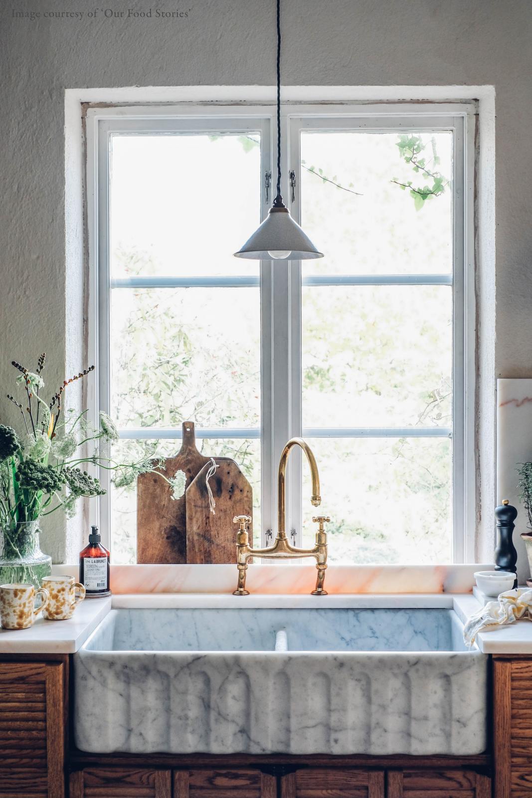 A closer look at a warm and inviting Haberdasher’s kitchen featuring a sink run with a Carrara marble worktop, a double-fluted marble sink, an aged brass Ionian tap, and prep tables with a copper worktop. Beautifully decorated with vintage items, artisan ceramics, wooden chopping boards, and a wild flower arrangement, this kitchen creates an airy and cosy Scandinavian small fishing town hideaway.