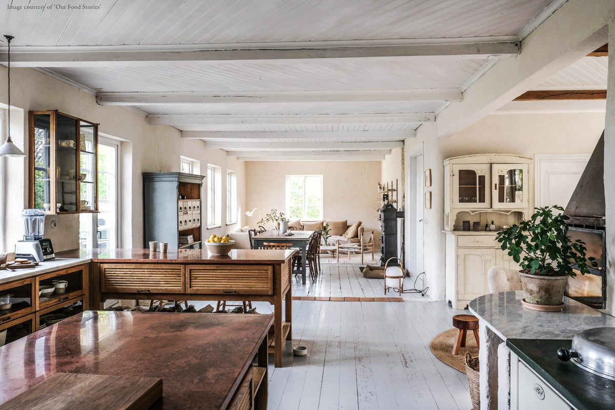 A warm and inviting Haberdasher’s kitchen features a sink and cooker run with a Carrara marble worktop, a double-fluted marble sink, an aged brass Ionian tap, prep tables with a copper worktop, and glazed wall cupboards. Beautifully decorated with vintage furniture and items, artisan ceramics, firewood, a small fireplace, and a wool armchair, this kitchen creates an airy and cosy Scandinavian small fishing town hideaway.