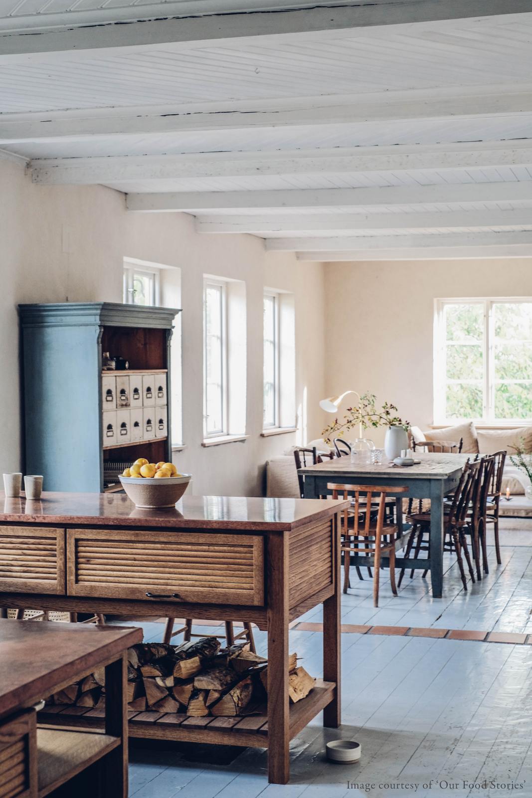 A warm and inviting Haberdasher’s kitchen features prep tables with a copper worktop. Beautifully decorated with vintage furniture and items, artisan ceramics, firewood, a bowl of lemons, a large couch, this space creates an airy and cosy Scandinavian small fishing town hideaway.
