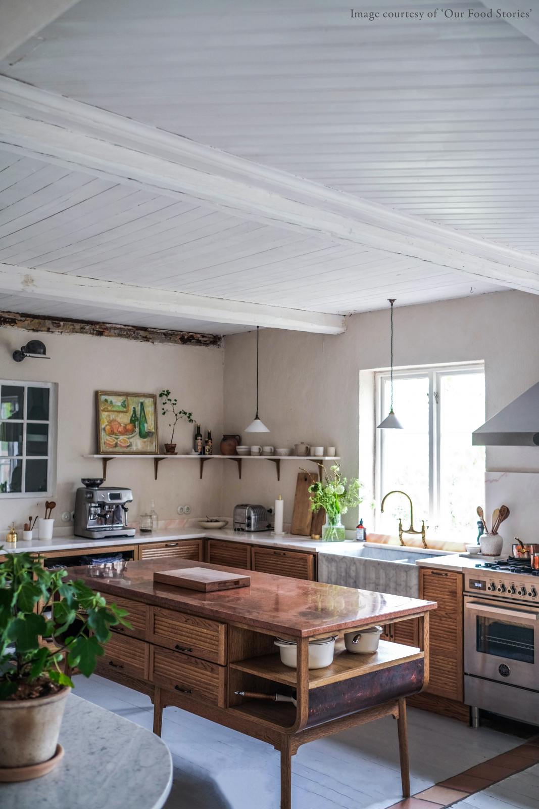An oblique view of a warm and inviting Haberdasher’s kitchen features a sink and cooker run with a Carrara marble worktop, a double-fluted marble sink, an aged brass Ionian tap, prep tables with a copper worktop, and a shelf. Beautifully decorated with vintage furniture and items, artisan ceramics, luscious green potted plants, and a still life painting, this kitchen creates an airy and cosy Scandinavian small fishing town hideaway.