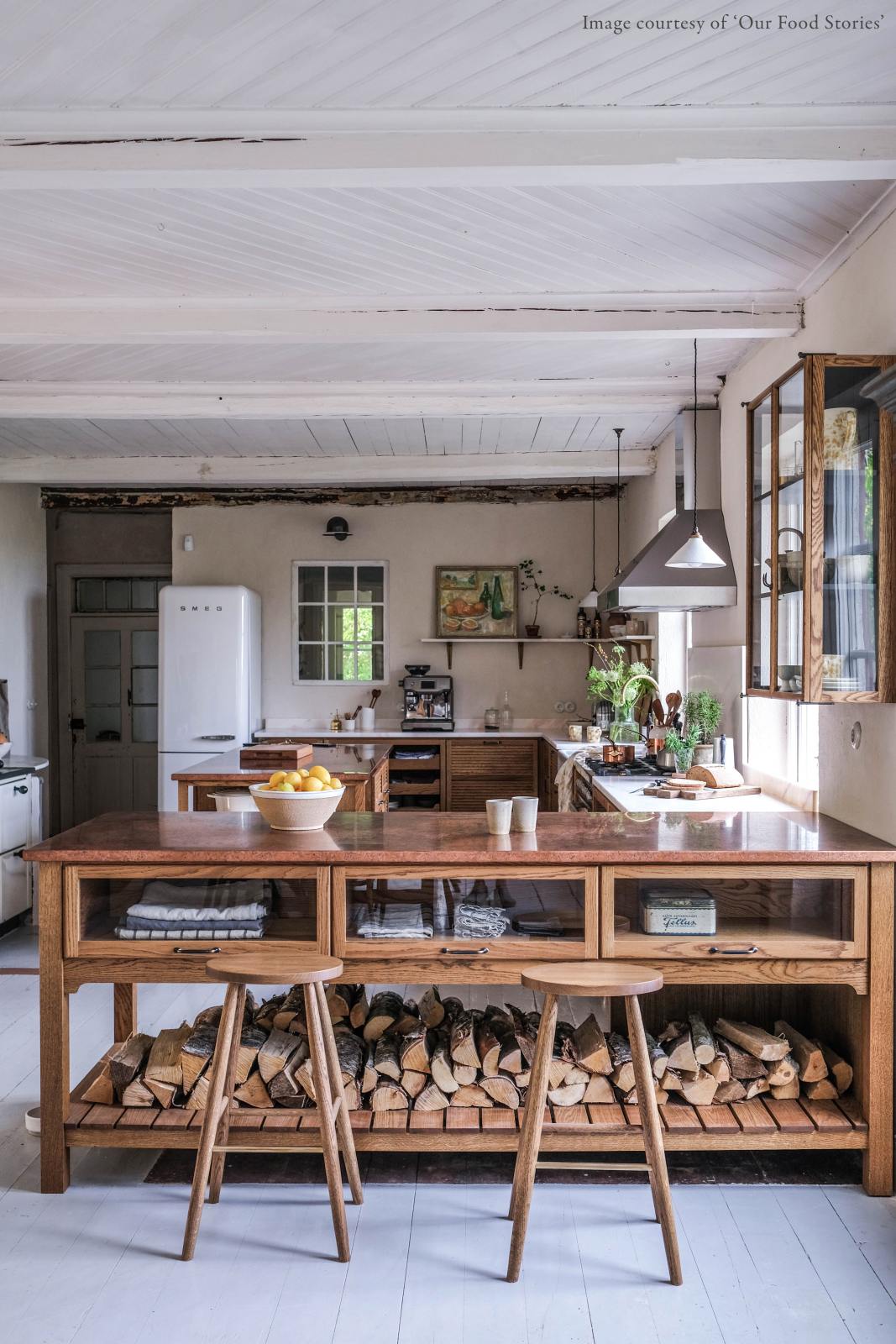 A warm and inviting Haberdasher’s kitchen features a sink and cooker run with a Carrara marble worktop, a double-fluted marble sink, an aged brass Ionian tap, prep tables with a copper worktop, glazed wall cupboards, and a shelf. Beautifully decorated with vintage furniture and items, artisan ceramics, firewood, Helen stools, and a bowl of lemons, this kitchen creates an airy and cosy Scandinavian small fishing town hideaway.