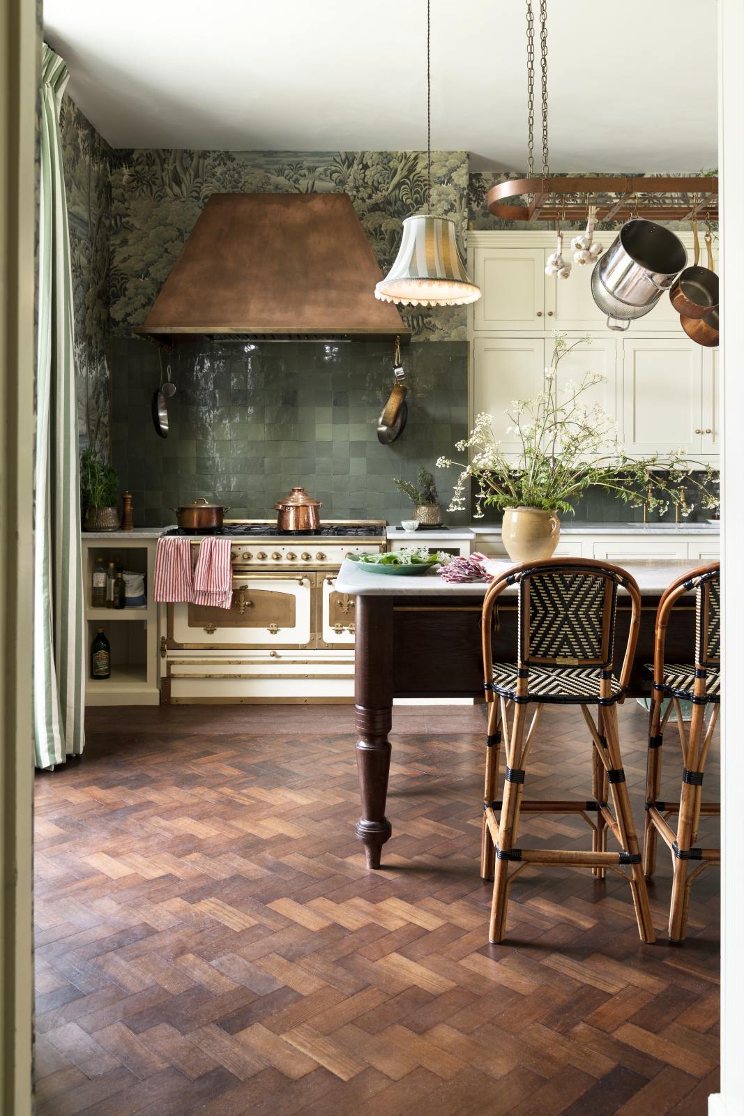A grand and eclectic kitchen features classic English cupboards with castellations, a Carrara marble worktop, and a stunning Heirloom dairy table. Beautifully decorated with detailed muted-green rainforest wallpaper, handmade Zellige tiles, a white and gold Italian gas cooker, a copper hood, and a white wildflower floral arrangement, this kitchen creates a fantasy experience in a castle.