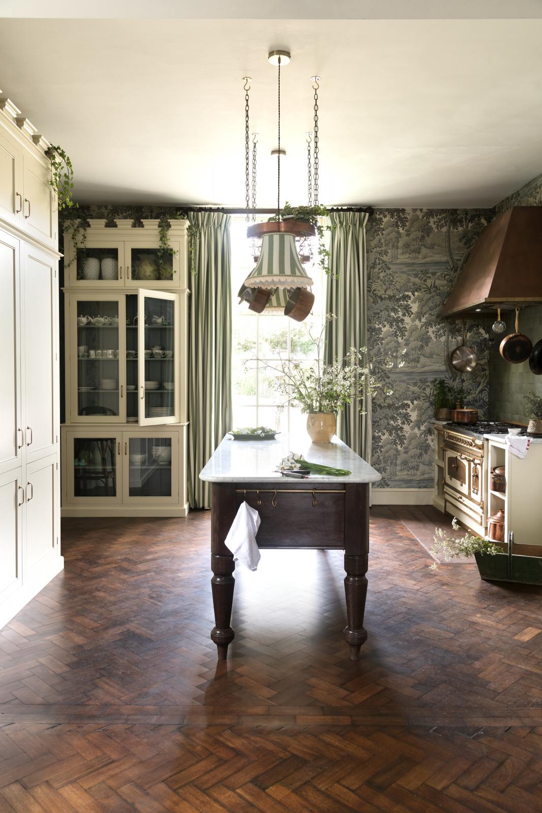 A grand and eclectic kitchen features classic English cupboards with castellations, a Carrara marble worktop, and a stunning Heirloom dairy table. Beautifully decorated with detailed muted-green rainforest wallpaper, handmade Zellige tiles, a white and gold Italian gas cooker, a copper hood, and a white wildflower floral arrangement, this kitchen creates a fantasy experience in a castle.