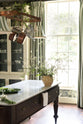 An oblique view of a grand and eclectic kitchen features classic English cupboards with castellations, a Carrara marble worktop, and a stunning Heirloom dairy table. Beautifully decorated with detailed muted-green rainforest wallpaper, artisan ceramics, copper pans, a green and white striped shade pendant light, and a white wildflower floral arrangement, this kitchen creates a fantasy experience in a castle.