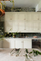 A grand and eclectic kitchen features a classic English sink run with a single copper sink, aged brass Mayan taps, wall cupboards, a Carrara marble worktop, and a stunning Heirloom dairy table. Beautifully decorated with detailed muted-green rainforest wallpaper, copper pans, a basket of fresh eggs, and a white wildflower floral arrangement, this kitchen creates a fantasy experience in a castle.