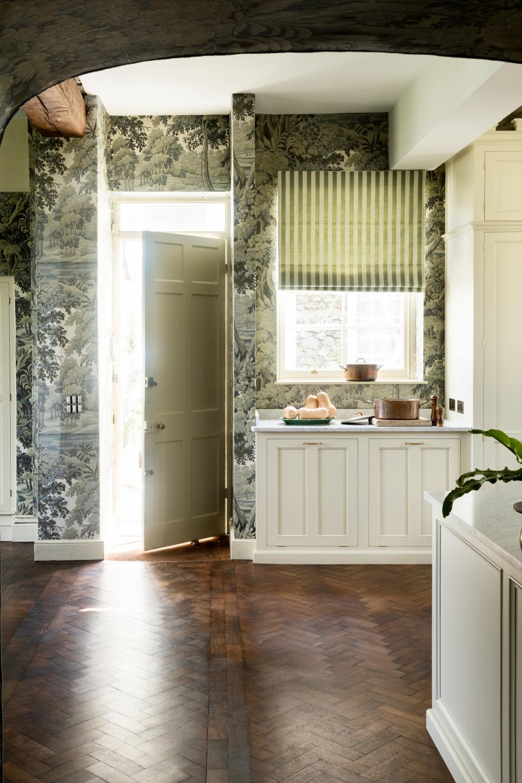 Past a dark archway is a grand and eclectic kitchen that features classic English cupboards with castellations, a Carrara marble worktop, and a stunning Heirloom dairy table. Beautifully decorated with detailed muted-green rainforest wallpaper, copper pans, a plate of butternut squash, a house plant peeking out, and the door to the outside slightly open, letting bright natural light in, this kitchen creates a fantasy experience in a castle.