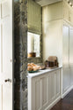 An oblique view of a grand and eclectic kitchen that features classic English cupboards with castellations, and a Carrara marble worktop. Beautifully decorated with detailed muted-green rainforest wallpaper, copper pans, a plate of butternut squash, and the door to the outside slightly open, letting bright natural light in, this kitchen creates a fantasy experience in a castle.