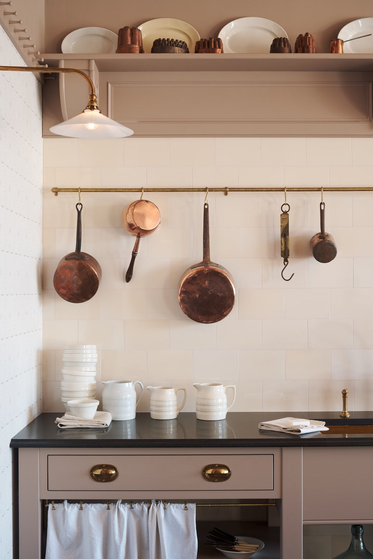 A quiet corner of a grand Princelet pink Heirloom and Classic English kitchen features a sink cabinet with a black granite worktop, aged brass rails, ditsy delft wallpaper, a tall cream subway tile splashback, and an Heirloom task light. Lovingly adorned with brass pots and pans and artisan ceramics, creating a warm, delicate, and airy kitchen space.
