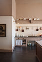 A quiet corner of a grand Princelet pink Heirloom and Classic English kitchen features a sink cabinet with a black granite worktop, a large kitchen island with a Carrara marble worktop, aged brass rails, ditsy delft wallpaper, a tall cream subway tile splashback, and an Heirloom task light. Lovingly adorned with brass pots and pans, an antique painting, and artisan ceramics, creating a warm, delicate, and airy kitchen space.