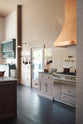 An oblique view of a quiet corner of a grand Princelet Pink Heirloom and Classic English kitchen features a cooker run with a black granite worktop, a large kitchen island with a Carrara marble worktop, an aged brass pot filler tap against a tall cream subway tile and border tile splashback, and a large copper canopy. Lovingly adorned with brass pots and pans, and artisan ceramics, creating a warm, delicate, and airy kitchen space.