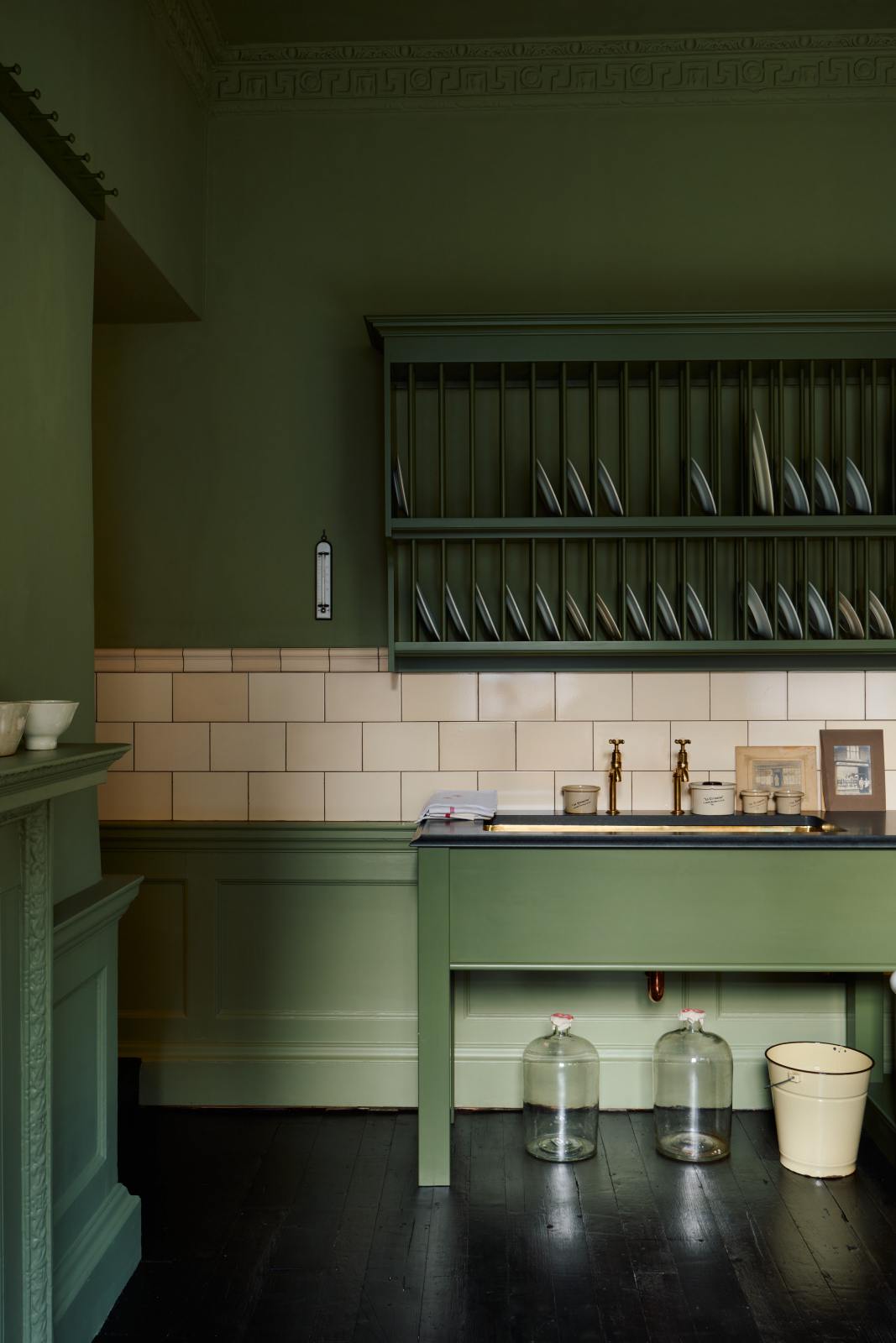 A quaint and stylish Wilkes green Heirloom kitchen features a sink cabinet with a café rail, white lace curtains, a polished brass sink, aged brass Mayan taps, and a large plate rack. Adorned with vintage photos, a cream bucket, simple ceramics, cream subway splashback tiles, and a golden-framed sliced green pumpkin, this kitchen creates a luxurious dining and social space.