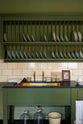 A quaint and stylish Wilkes green Heirloom kitchen features a sink cabinet with a café rail, white lace curtains, a polished brass sink, aged brass Mayan taps, and a large plate rack. Adorned with vintage photos, a cream bucket, simple ceramics, cream subway splashback tiles, and a golden-framed sliced green pumpkin, this kitchen creates a luxurious dining and social space.