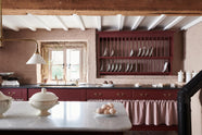 A refectory-red Heirloom kitchen’s sink cabinet reveals a polished brass sink, aged brass Mayan taps, café curtain rails, a red and white striped curtain, a plate rack, and an Heirloom dairy table. Beautifully decorated with Heirloom gaselier lights, a vintage scale, artisan ceramics, and a plate of fresh eggs, this kitchen offers a modern twist on vintage furniture, creating a classic range designed to last.