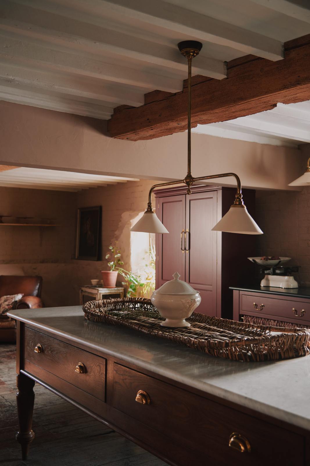 An oblique view of a refectory-red Heirloom kitchen features a sink cabinet with café curtain rails, a red and white striped curtain, a dairy table, and a pantry cupboard. Beautifully decorated with Heirloom gaselier lights, a vintage scale, artisan ceramics, and a large woven placemat, this kitchen offers a modern twist on vintage furniture, creating a classic range designed to last.