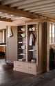 A pink freestanding pantry cupboard situated in the berry-red Heirloom kitchen at Cotes Mill. The double doors are open to show 5 shelves on the left, housing linens and crockery, and on the right there are just two shelves, with most of the space reserved for an aged brass hanging rail. The base of the pantry is made of two drawers which protrude slightly to make a step or seat when the doors are closed.