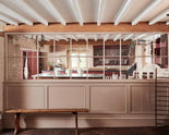 Past a princelet-pink glazed partition is a refectory-red Heirloom kitchen featuring a sink cabinet with a polished brass sink, aged brass Mayan taps, café curtain rails, a red and white striped curtain, a dairy table, a cooker run, a vegetable box, and a plate rack.