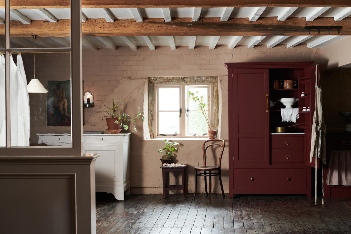 A quiet corner of a refectory-red Heirloom kitchen features a pantry cupboard. Beautifully decorated with vintage furniture, house plants, a creamware pendant light, and an aged brass café curtain rail, this kitchen offers a modern twist on vintage furniture, creating a classic space designed to last.